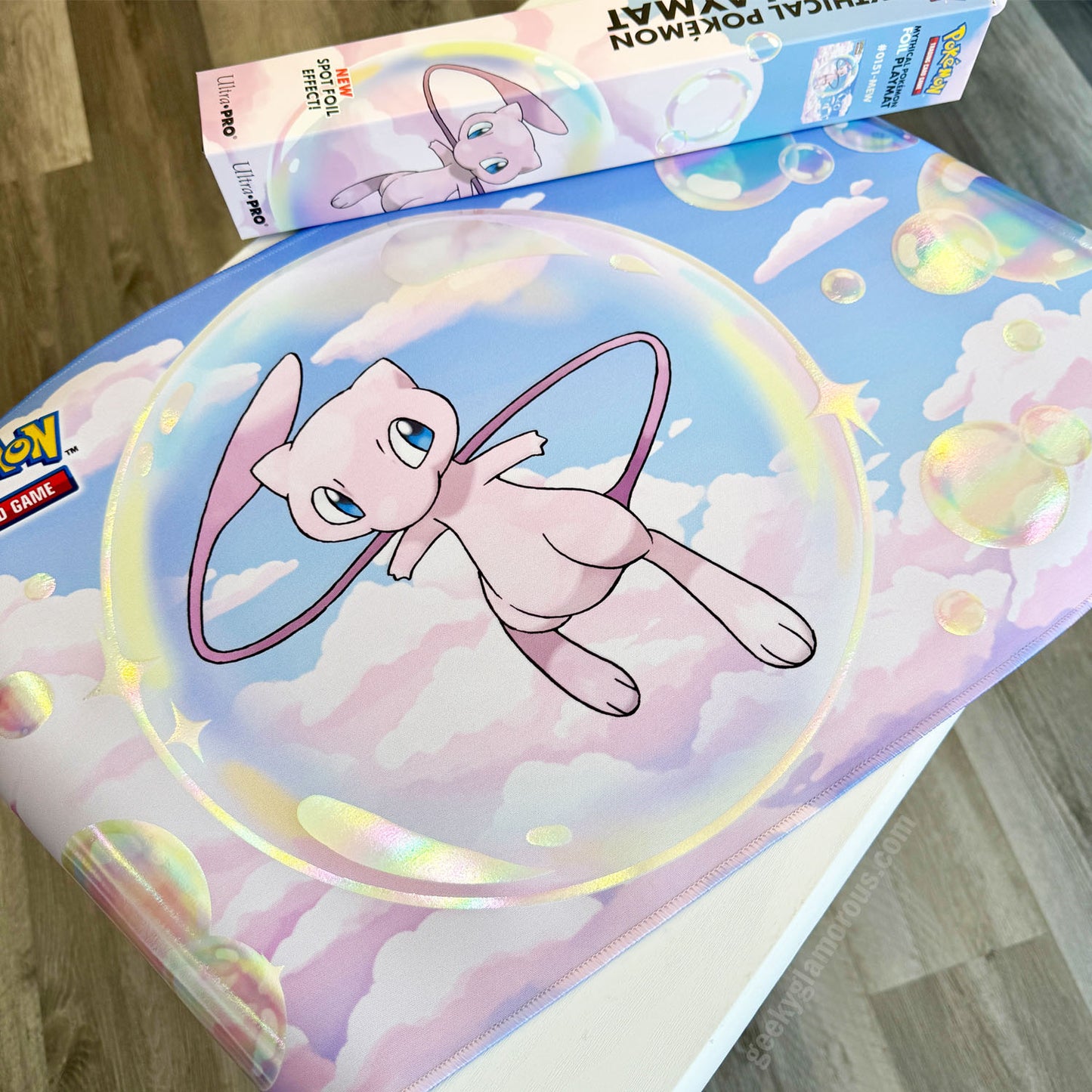 A Pokémon-themed playmat featuring Mew floating in a bubble among pastel clouds, with a matching Pokémon playmat box partially visible behind it on a wooden floor.
