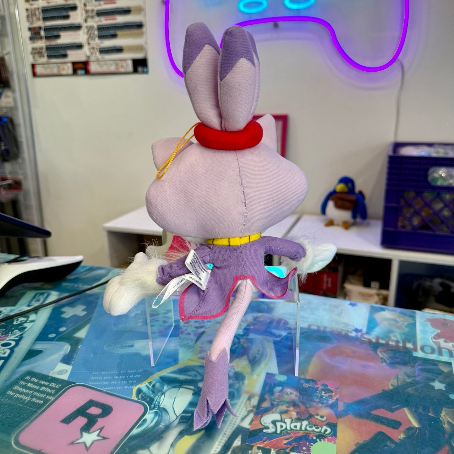 A plush toy of a purple cartoon character with cat ears, a red headband, and a purple outfit, shown from the back on clear display stands in a room setting.