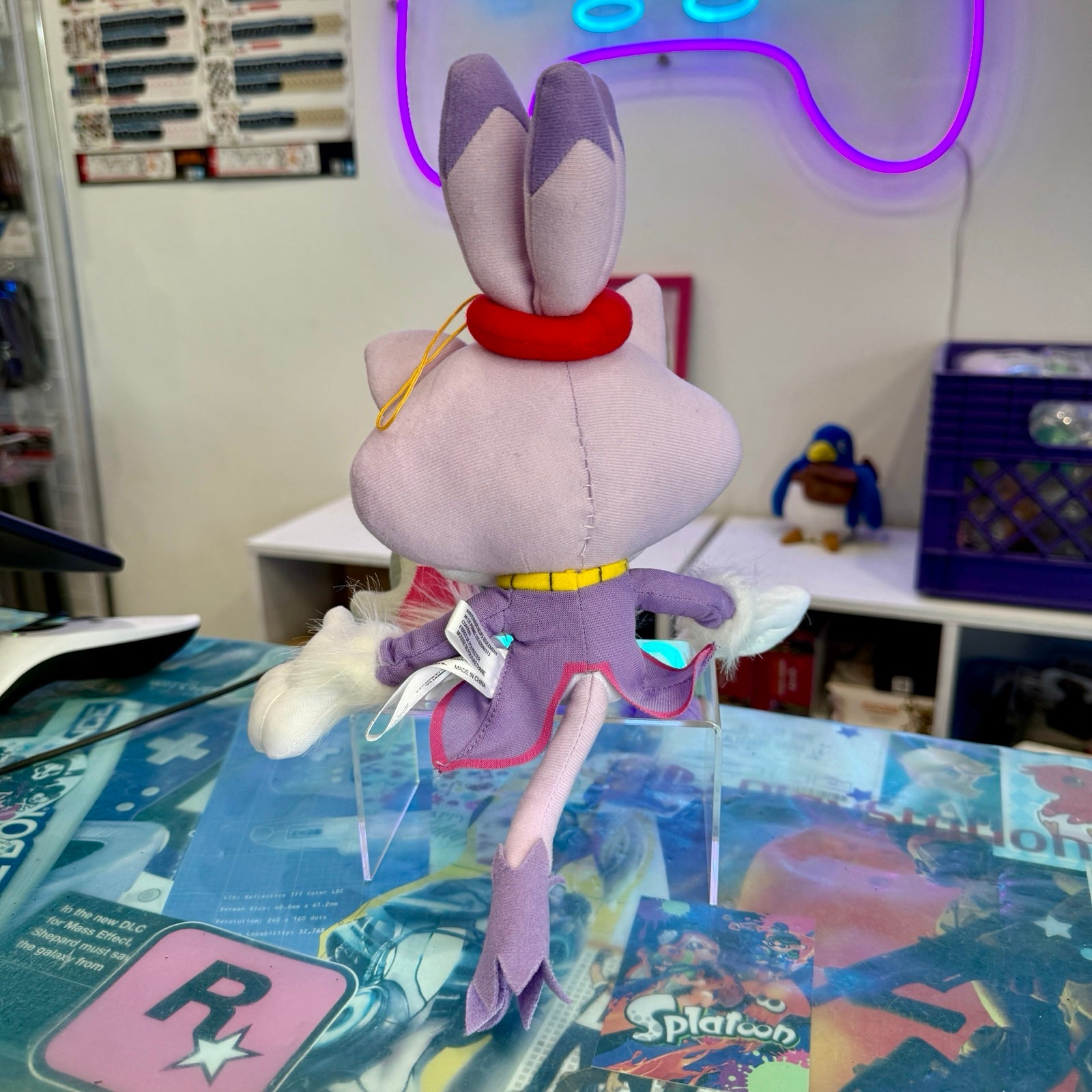 A plush toy of a purple cartoon character with cat ears, a red headband, and a purple outfit, shown from the back on clear display stands in a room setting.