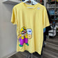 A yellow short-sleeve graphic T-shirt hanging on a white hanger, featuring a cartoon illustration of Wario. The shirt is displayed in a retail store setting.