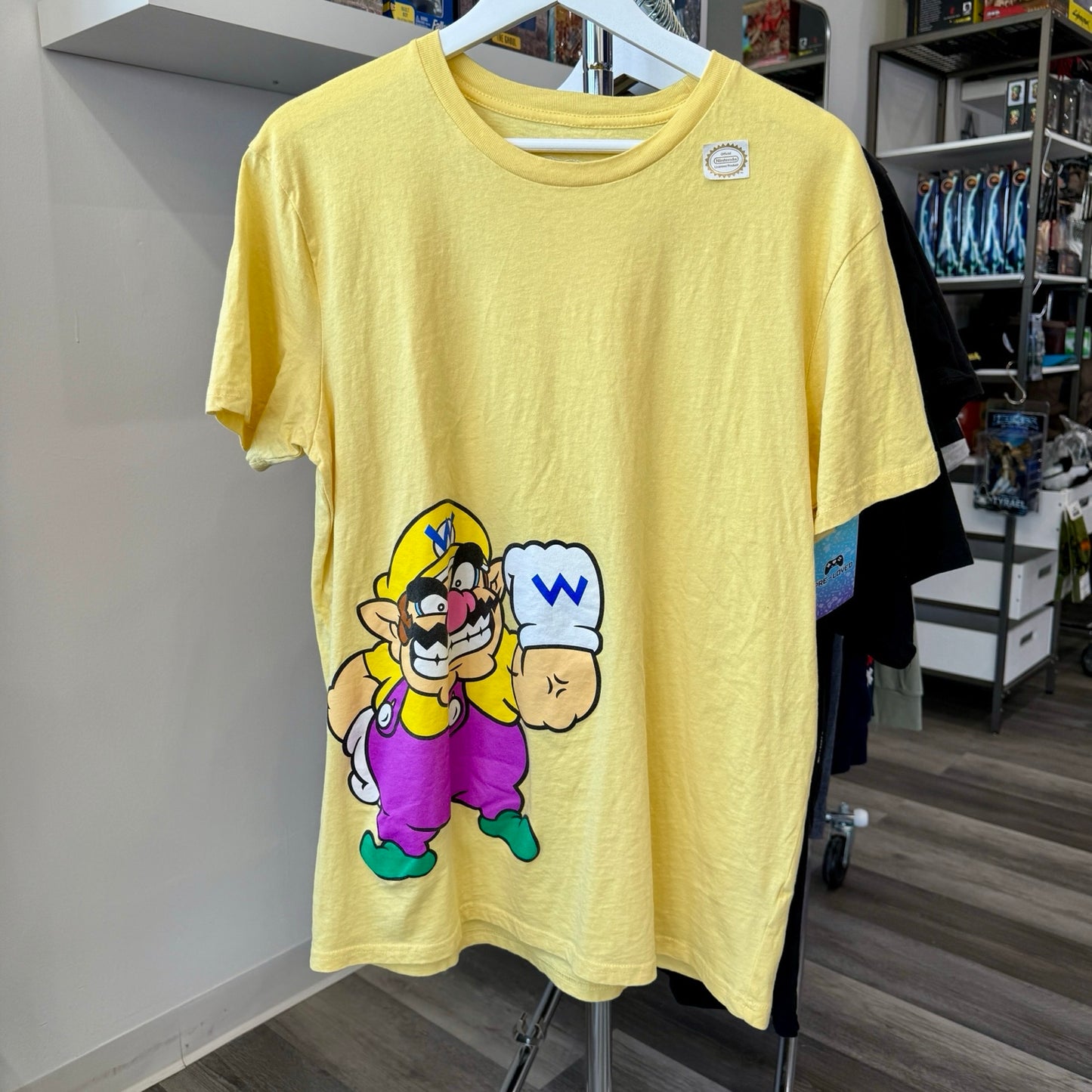 A yellow short-sleeve graphic T-shirt hanging on a white hanger, featuring a cartoon illustration of Wario. The shirt is displayed in a retail store setting.