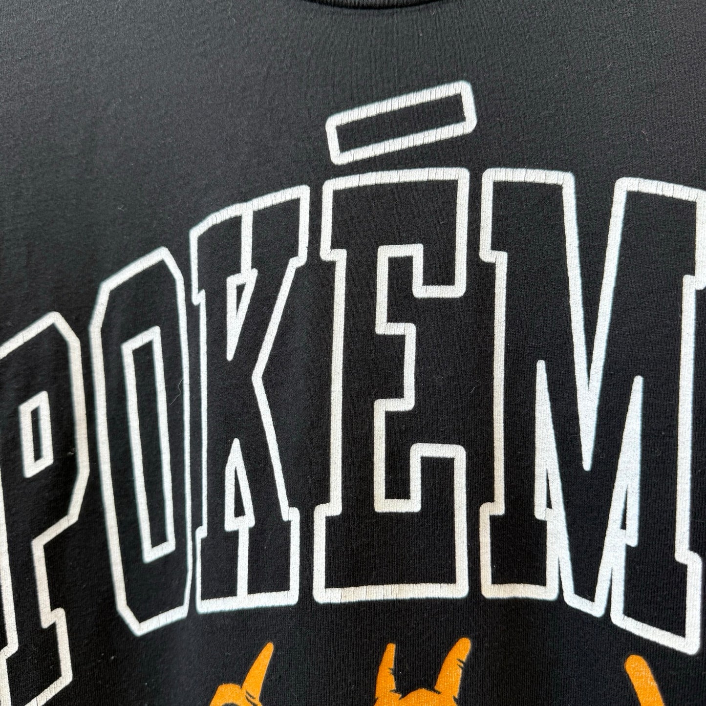 Close-up of a black T-shirt featuring large white outlined POKEMON lettering and orange graphic elements below.