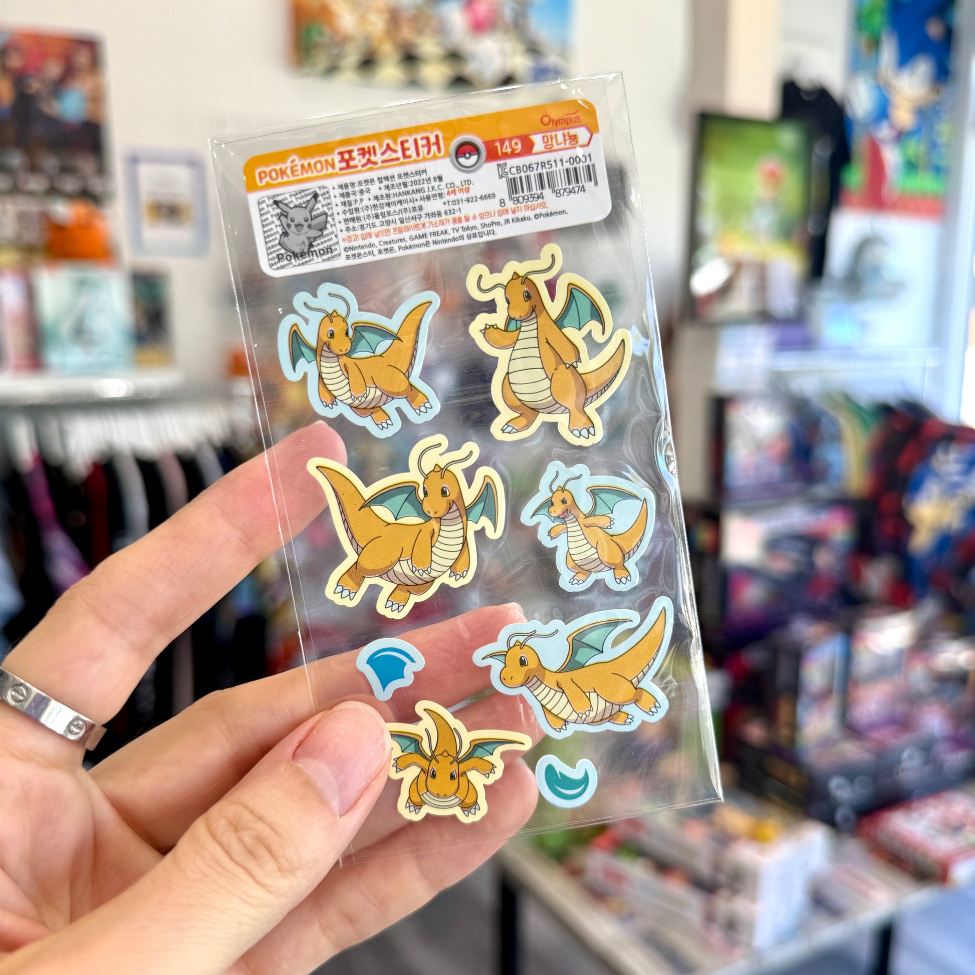 A hand holds a clear plastic package of six Dragonite Pokémon stickers in a retail store setting with shelves and products blurred in the background.
