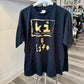 A black short-sleeve graphic t-shirt hanging on a white hanger on a clothing rack inside a retail store. The shirt features a distressed gold graphic print on the front with the text 'ki' inside a rough square, 'killer instinct' underneath, and '4 life' below.