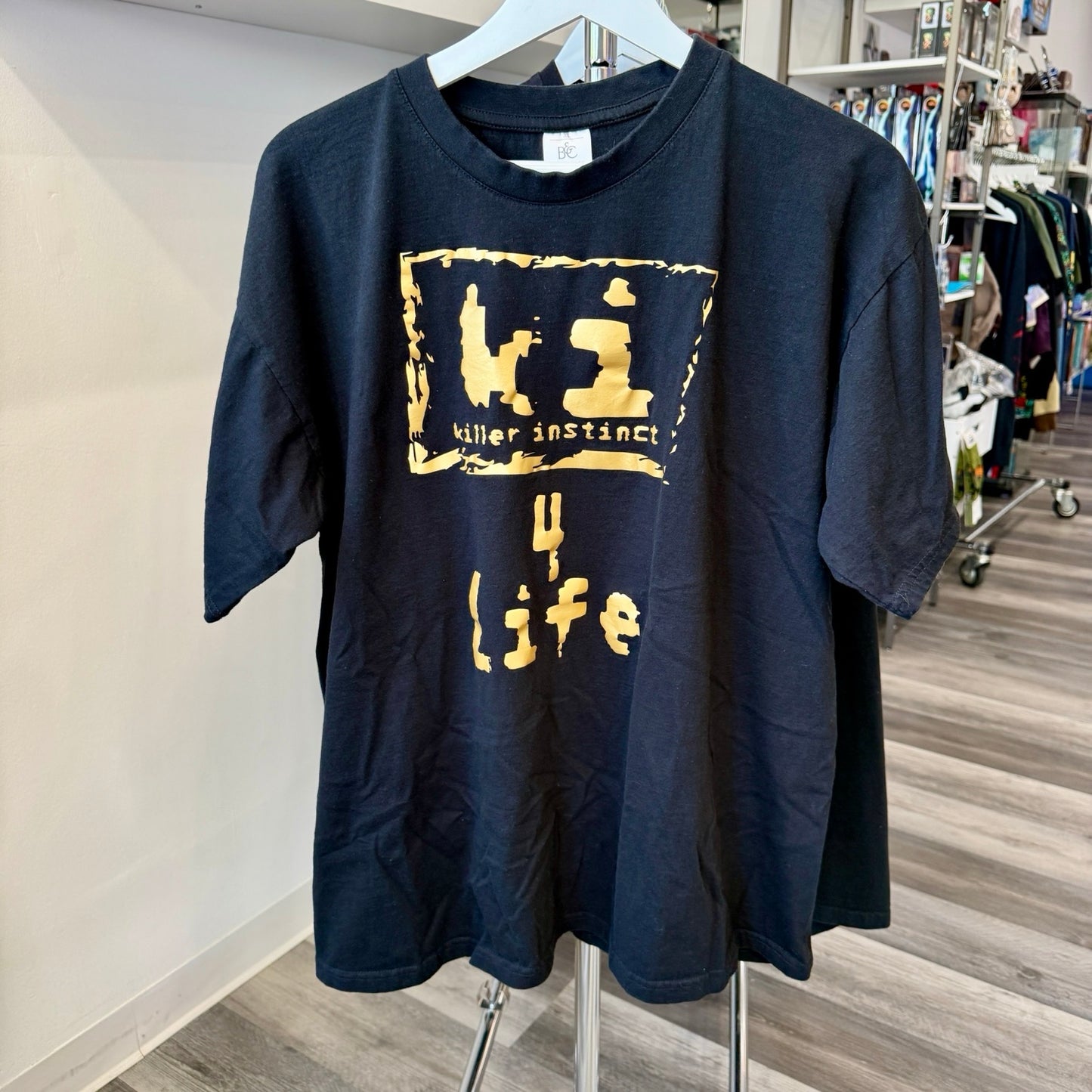 A black short-sleeve graphic t-shirt hanging on a white hanger on a clothing rack inside a retail store. The shirt features a distressed gold graphic print on the front with the text 'ki' inside a rough square, 'killer instinct' underneath, and '4 life' below.