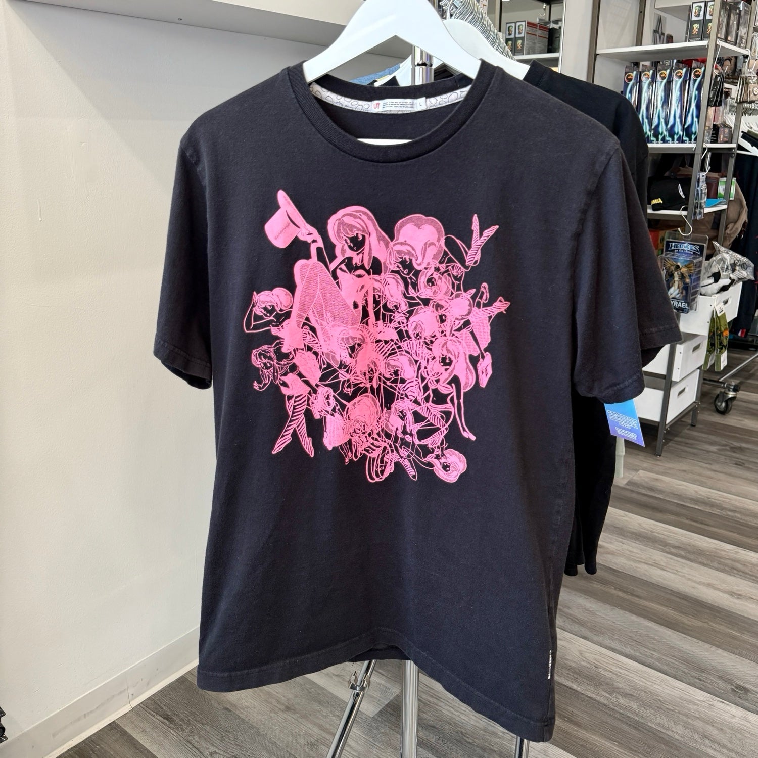 A black short-sleeve graphic t-shirt hanging on a white hanger, featuring a large pink anime-style character print on the front. The shirt is displayed on a clothing stand inside a retail store.