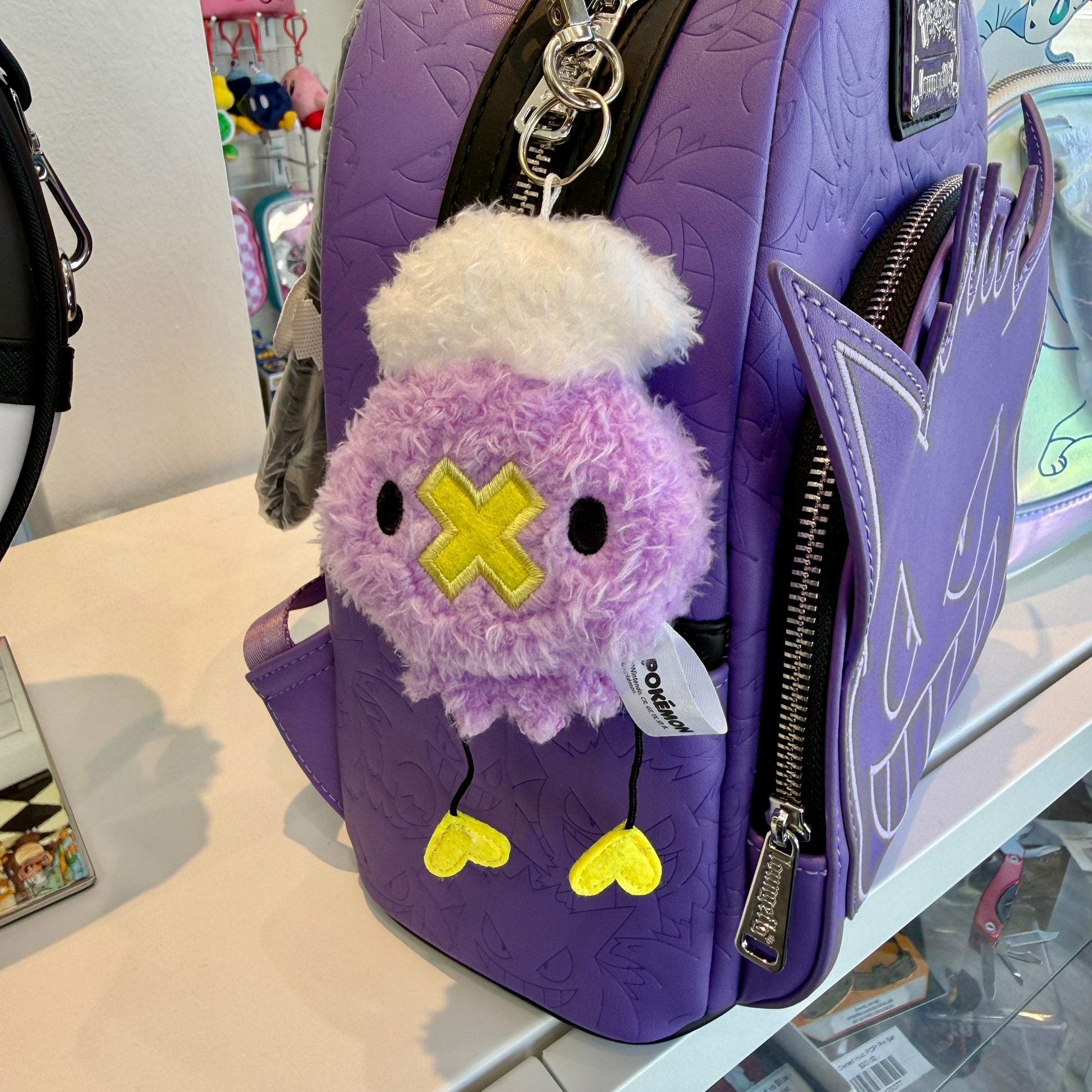 Fluffy keychain charm of the Pokemon, Drifloon hanging off a purple backpack in a store. 