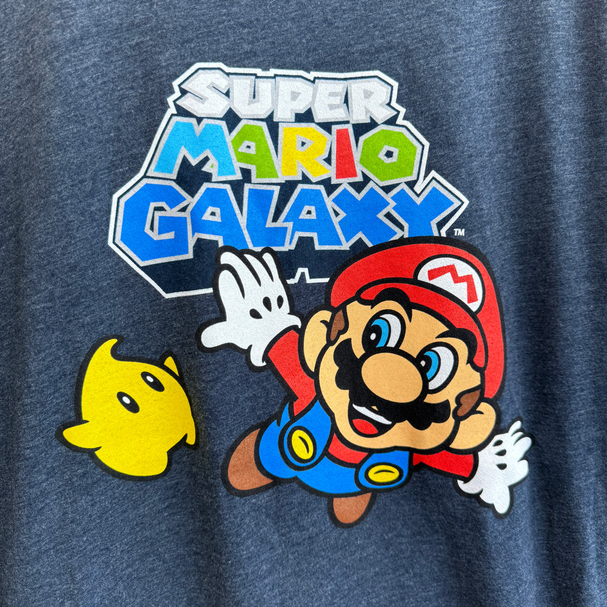 Close-up of a heather navy blue t-shirt featuring a large graphic print of the 'Super Mario Galaxy' logo and characters Mario and a yellow star.