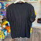 A black short-sleeve t-shirt hanging on a white hanger in a retail store, showing the plain back of the garment. The background contains shelves with various colorful plush toys and store fixtures.