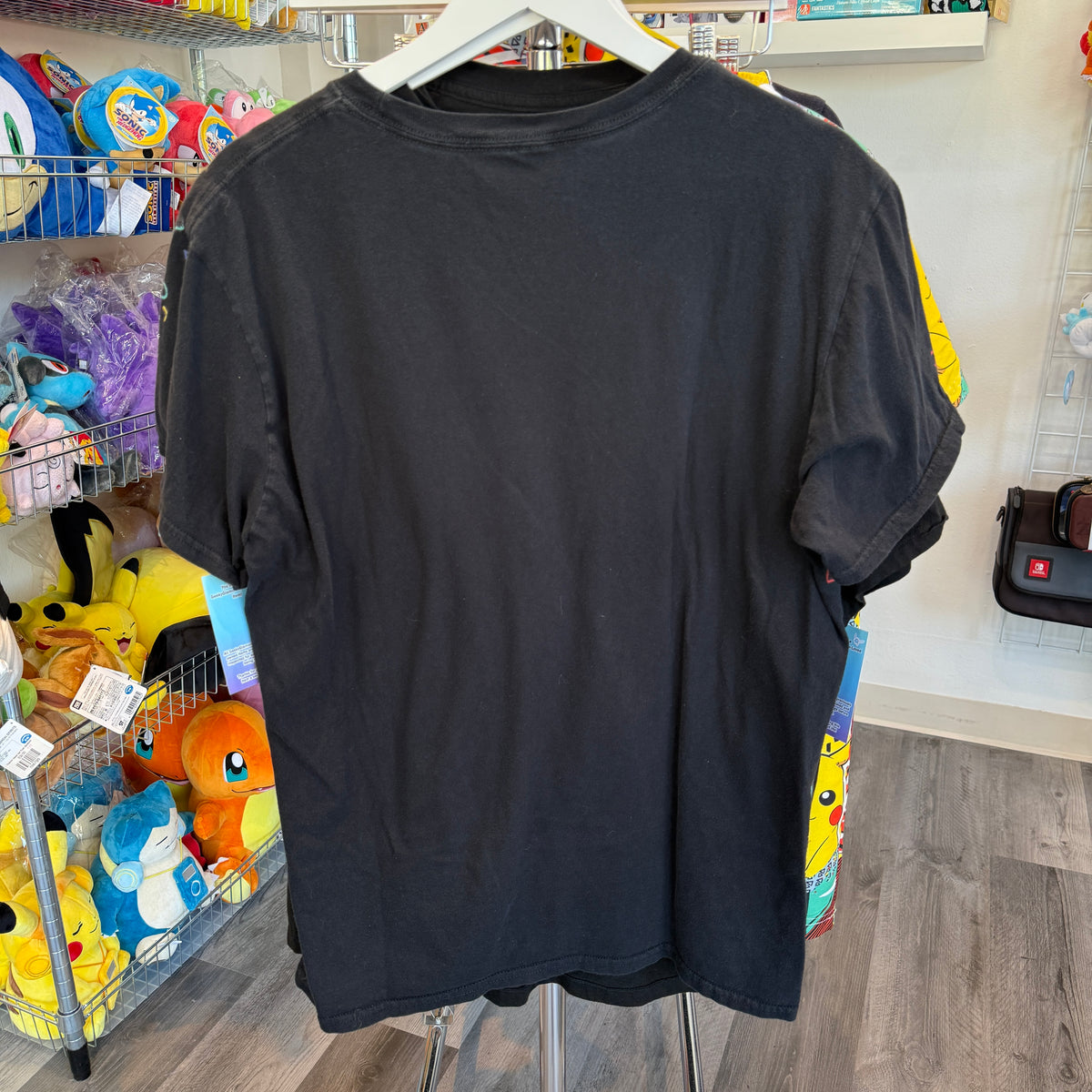 A black short-sleeve t-shirt hanging on a white hanger in a retail store, showing the plain back of the garment. The background contains shelves with various colorful plush toys and store fixtures.