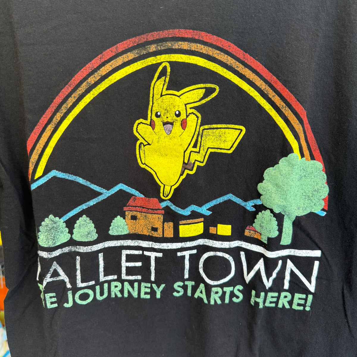 Close-up of a black graphic T-shirt featuring Pikachu flying over a rainbow and mountain scene with the text 'Pallet Town' and 'The Journey Starts Here!'.