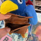 A close-up of a brown faux-leather coin purse being held open by a hand, attached to a blue and yellow plush penguin toy. The background shows a busy retail store shelf.