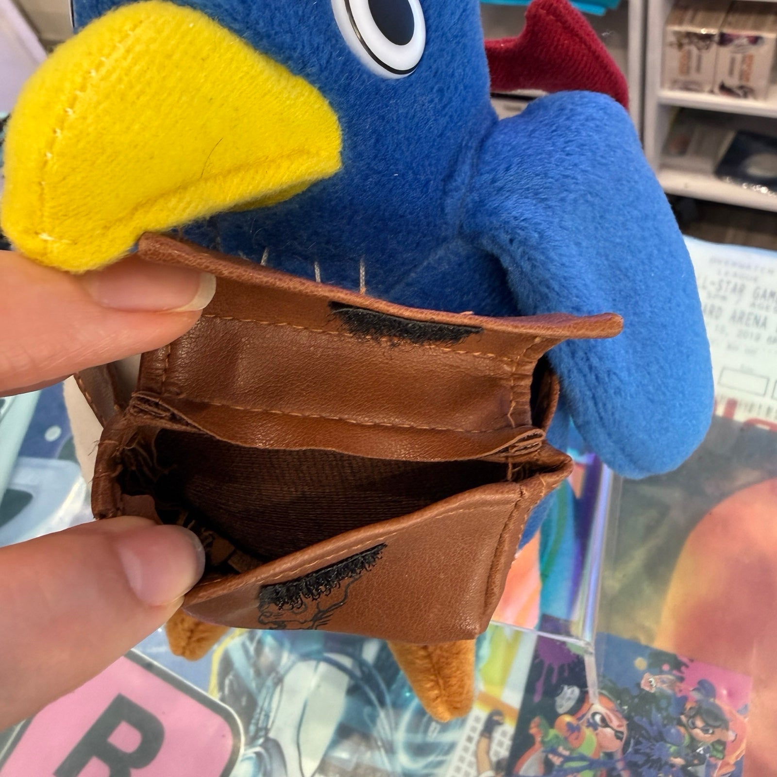 A close-up of a brown faux-leather coin purse being held open by a hand, attached to a blue and yellow plush penguin toy. The background shows a busy retail store shelf.