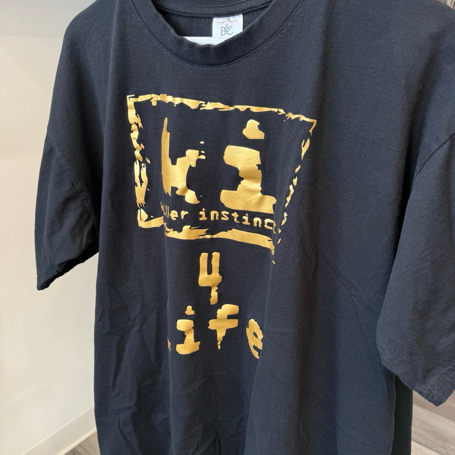 A dark charcoal gray t-shirt with a distressed gold graphic print on the front, featuring the text '4 life' and a stylized symbol. The shirt is shown hanging against a plain indoor wall.