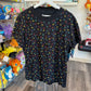 A black short-sleeve t-shirt with a colorful all-over pattern of PlayStation controller-style symbols (circles, squares, triangles, crosses) hanging on a white hanger in a retail store.