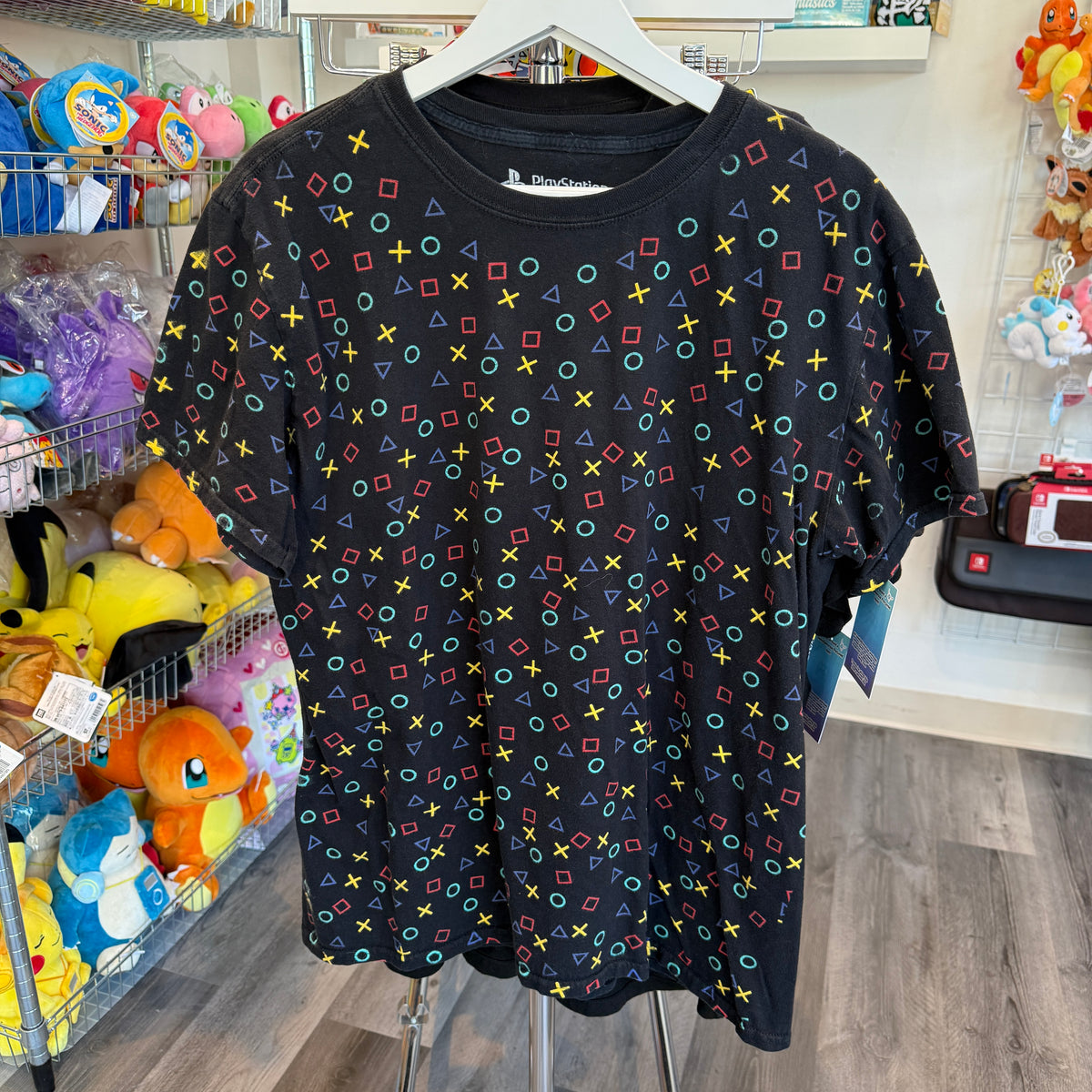 A black short-sleeve t-shirt with a colorful all-over pattern of PlayStation controller-style symbols (circles, squares, triangles, crosses) hanging on a white hanger in a retail store.