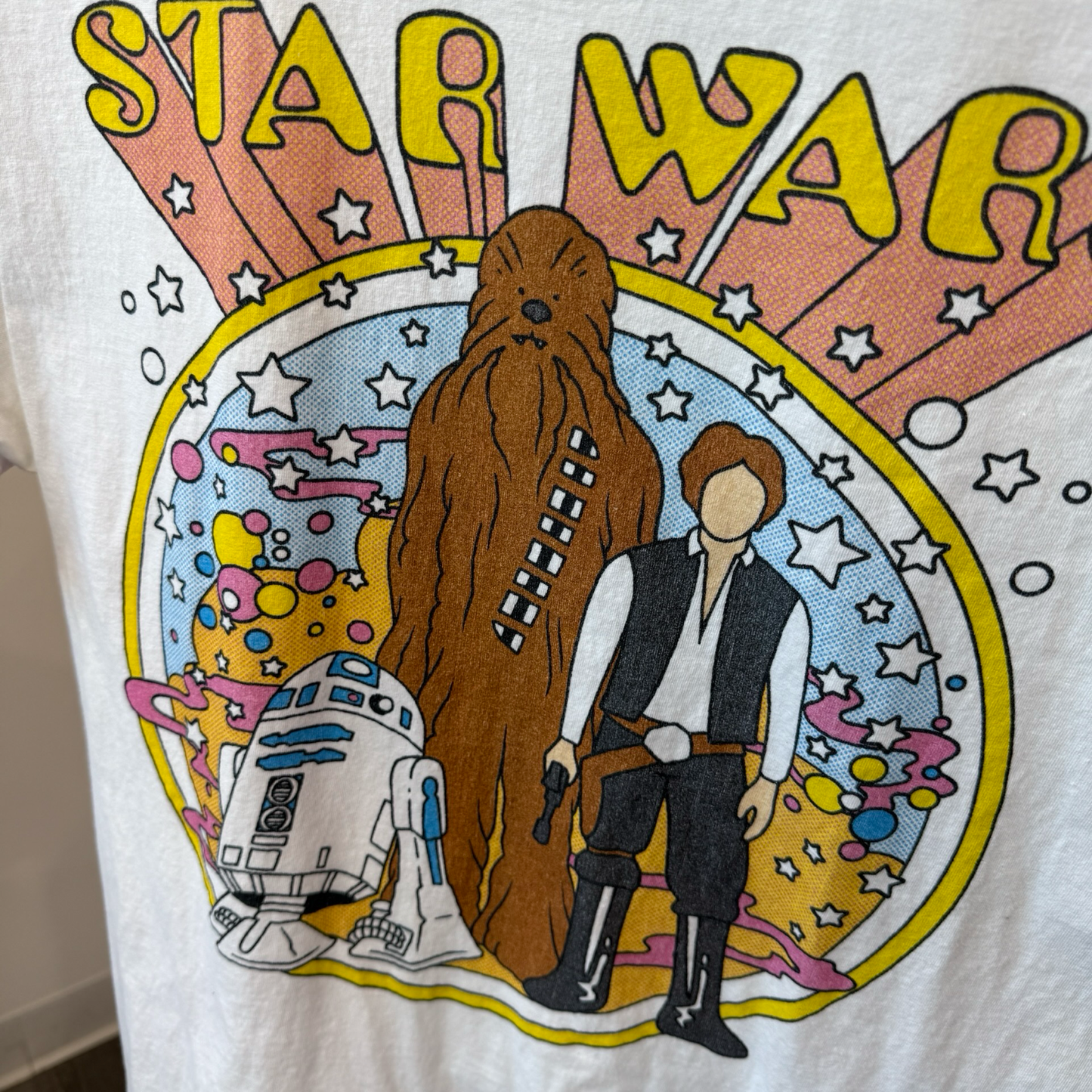 Close up shot of white tshirt with 1960s inspired graphic of R2D2, Chewbacca, and Han Solo from Star Wars. 
