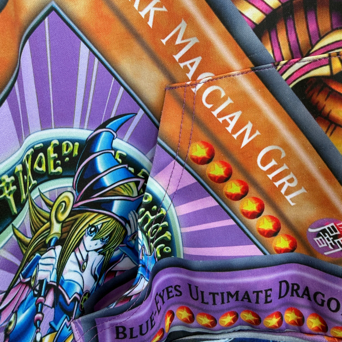 Close-up of a colorful printed textile featuring anime-style artwork, including a blue wizard character, purple and orange diagonal bands, and text such as 'MAGICIAN GIRL' and 'BLUE EYES ULTIMATE DRAGON'.