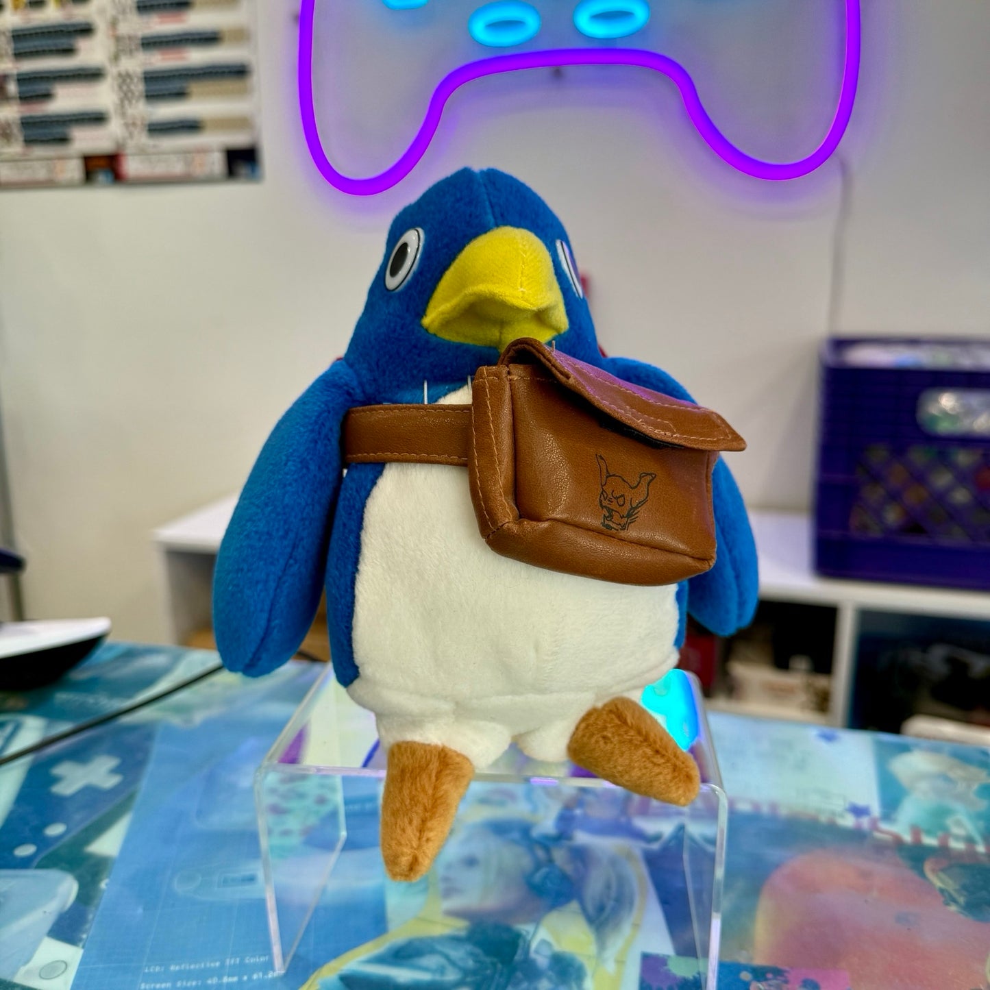 A blue and white penguin plush toy wearing a brown leather-look pouch with a fox logo, displayed on a clear acrylic stand on a desk with a blurred room background.