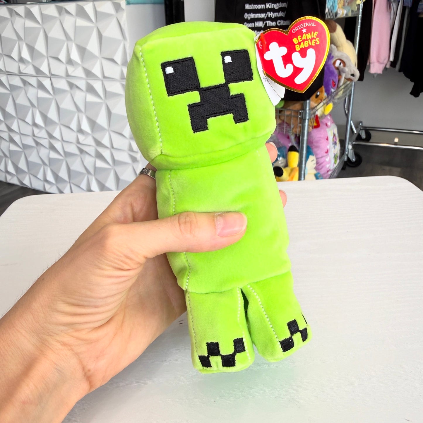 A hand holding a bright green Minecraft Creeper plush toy with a red Ty Beanie Babies hangtag, set against a store background.