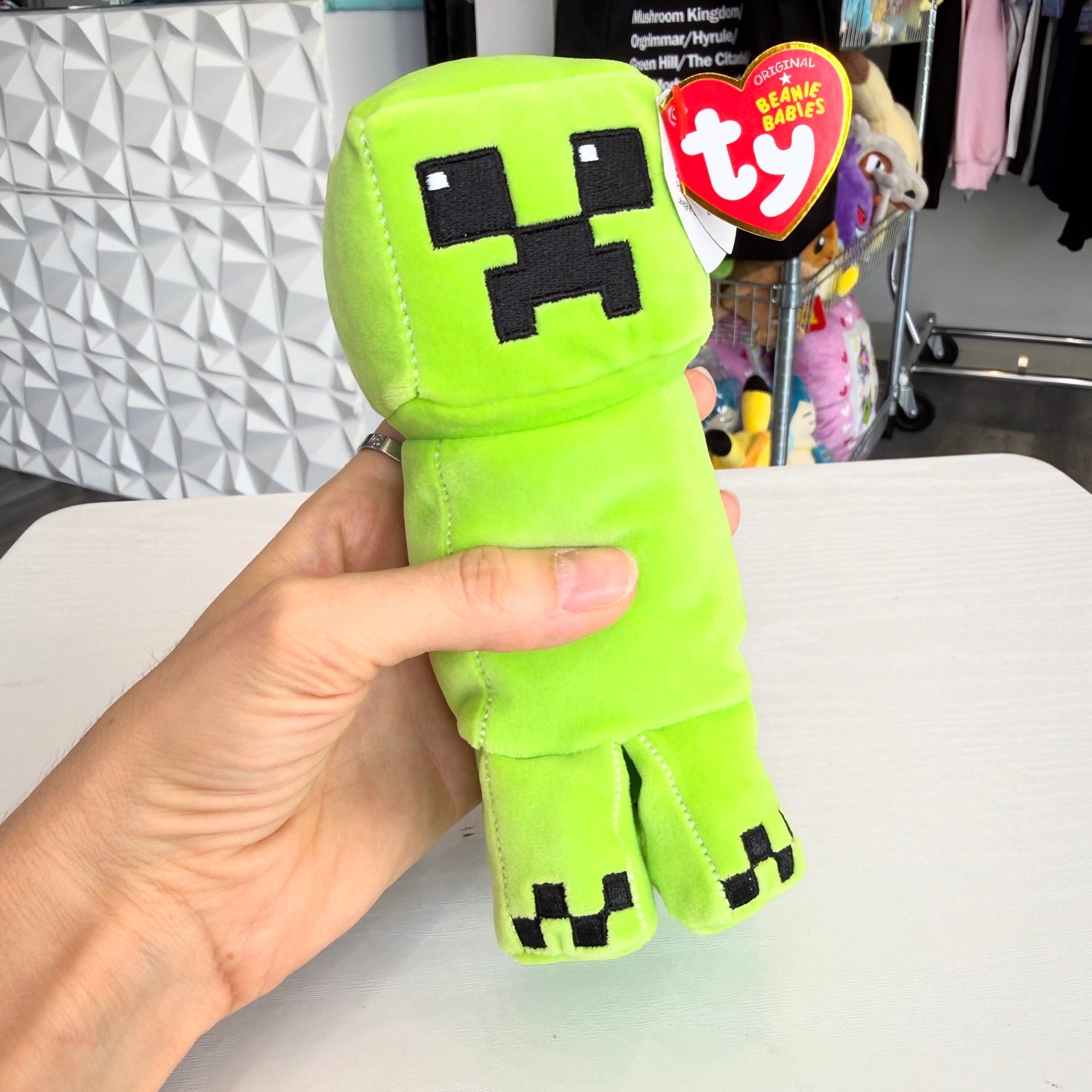 A hand holding a bright green Minecraft Creeper plush toy with a red Ty Beanie Babies hangtag, set against a store background.