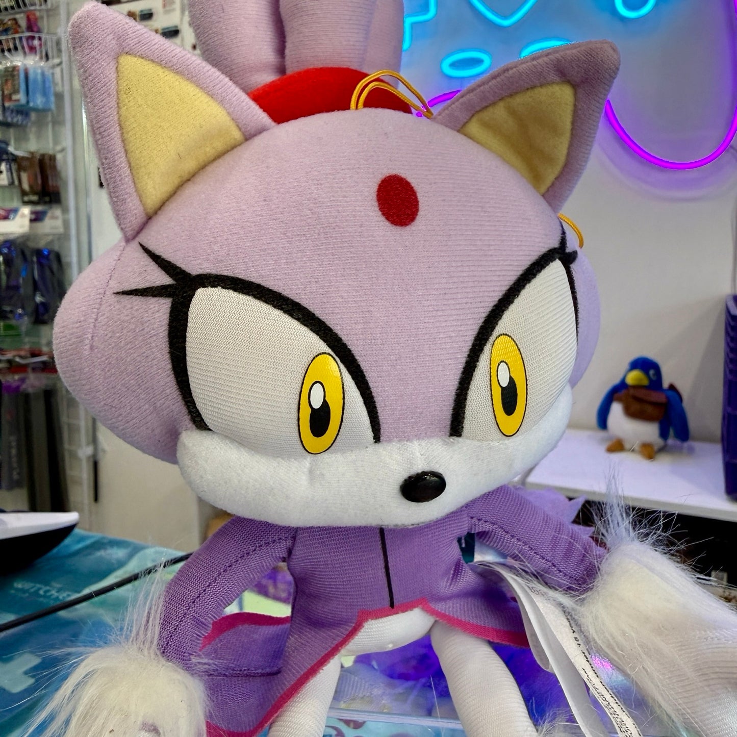 A plush toy of a purple cat character with yellow eyes, a red forehead emblem, and a purple outfit, photographed in a retail or studio setting with colorful neon lights in the background.