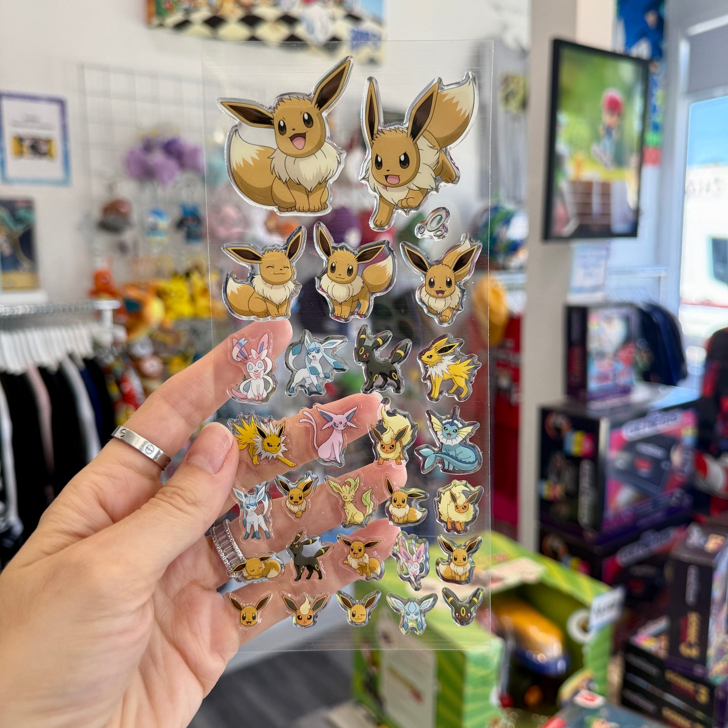A hand holds a clear plastic sheet covered with multiple small Eevee character stickers in different colors, photographed inside a retail store with shelves and merchandise blurred in the background.