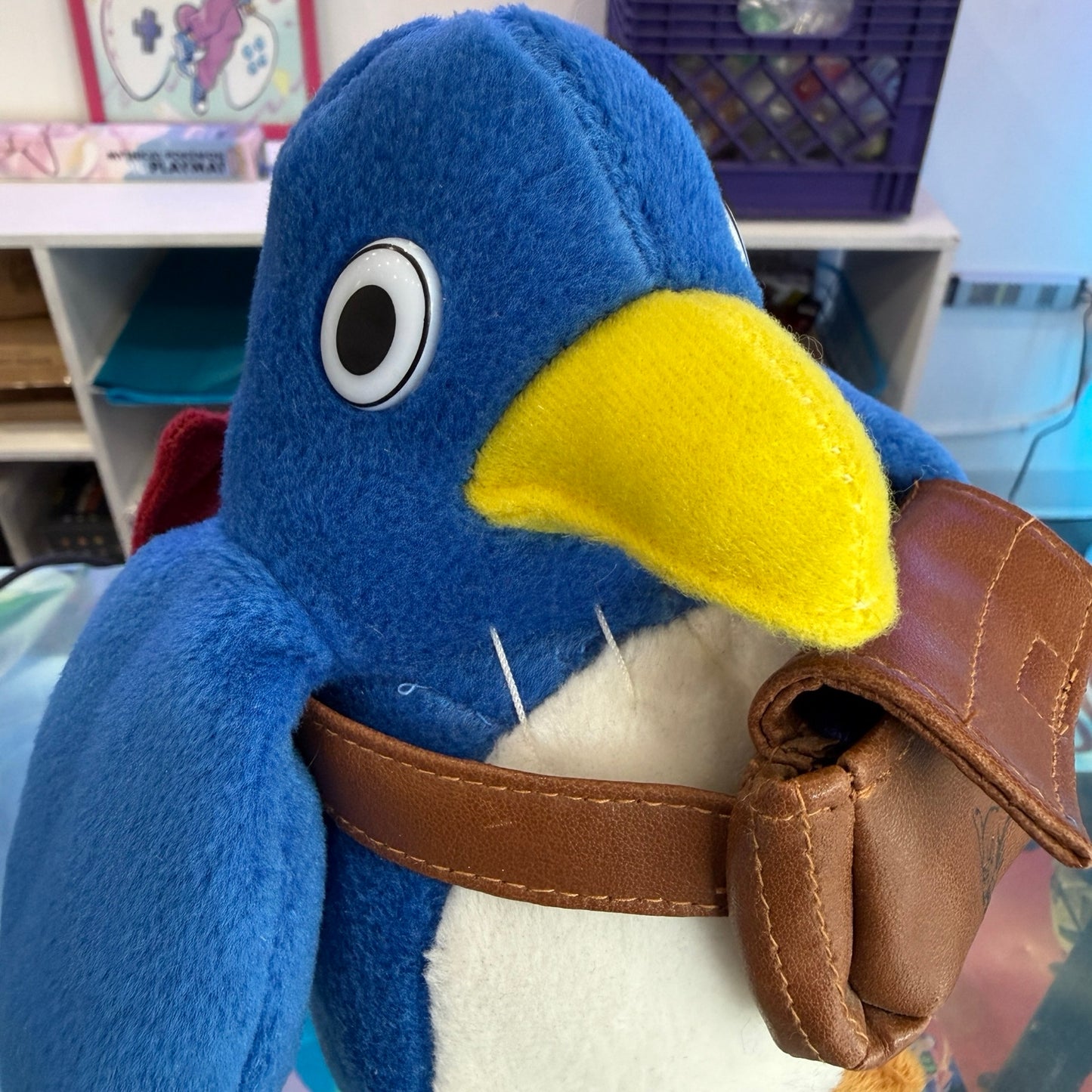A blue penguin plush toy with a yellow beak, white belly, brown leather-like harness, and brown glove, photographed indoors with shelves and toys in the background.