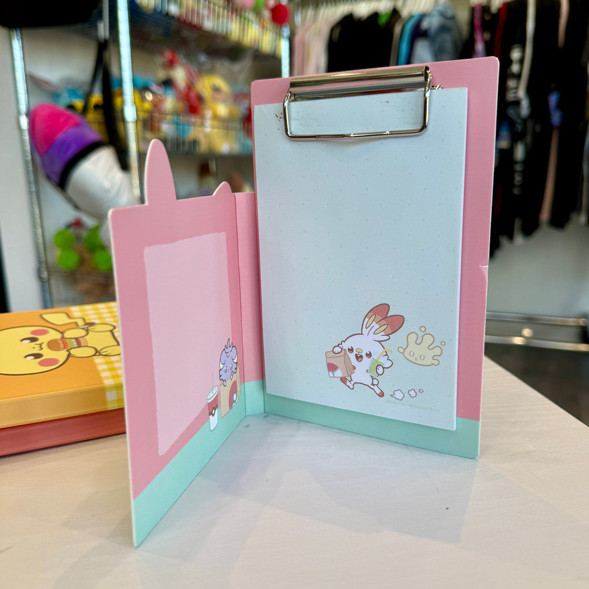 A pink and green Pokémon themed memo pad clipboard with a store in the background. 