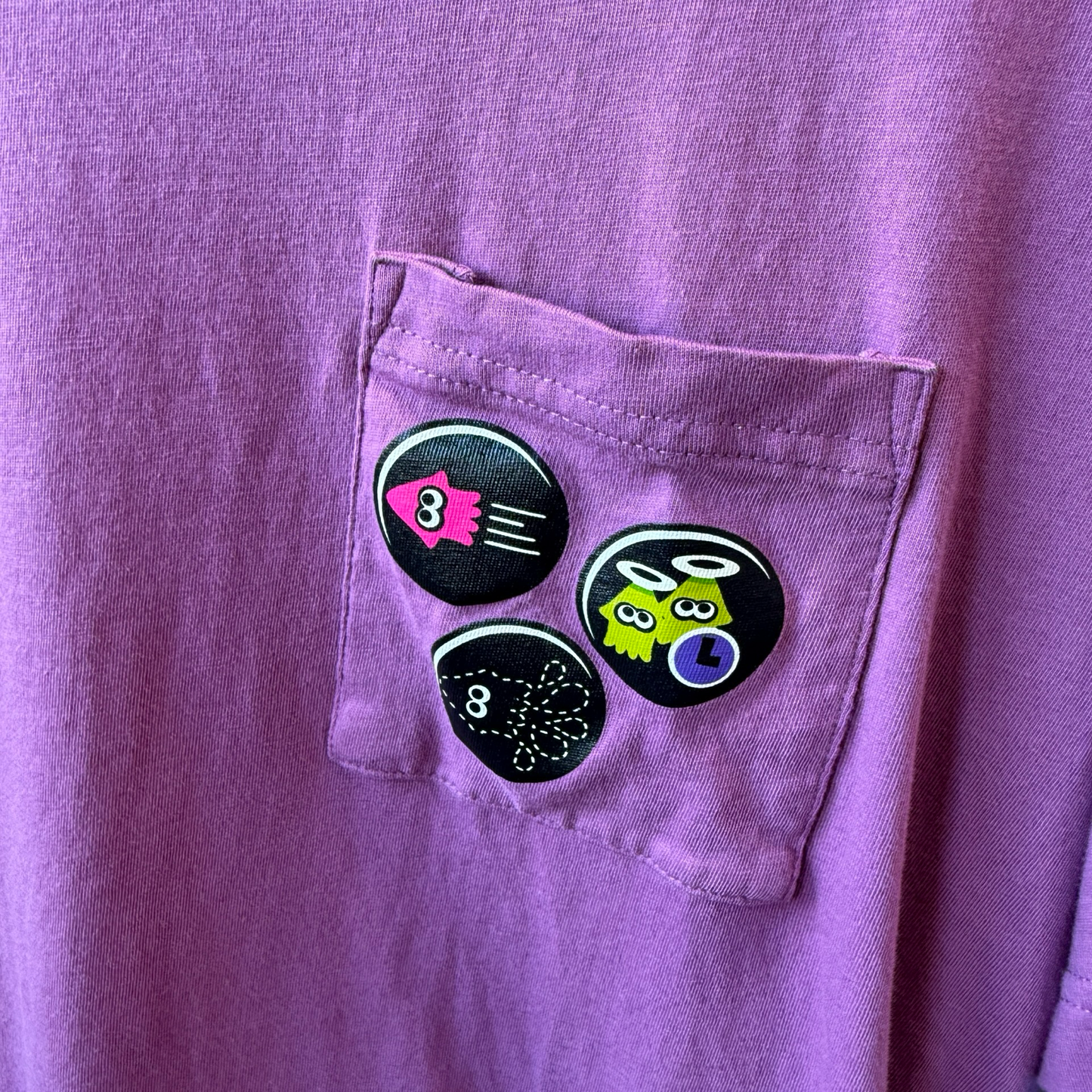 Up close view of purple shirt with pocket. On the pocket are three circular graphics of squids from the video game, Splatoon. 