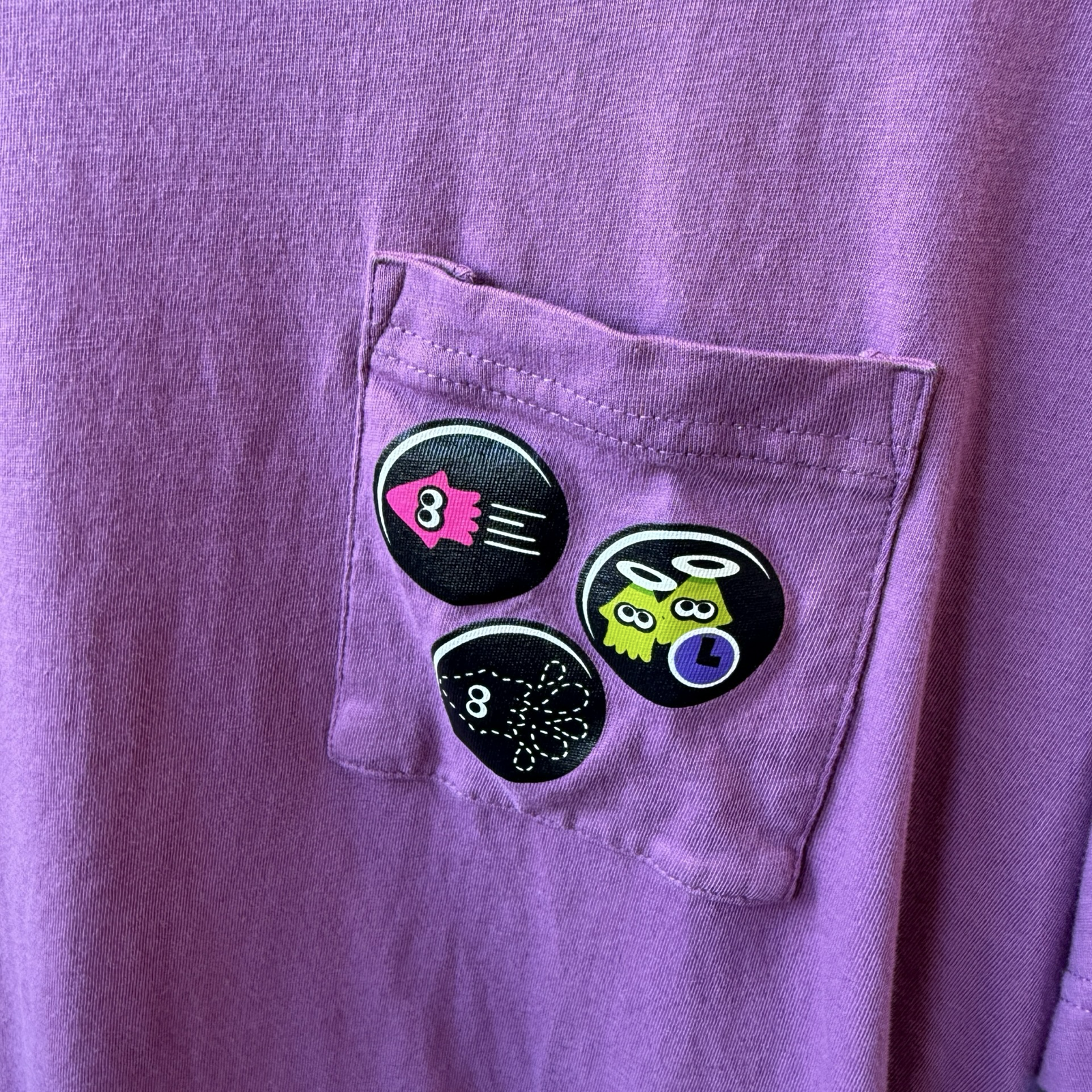 Up close view of purple shirt with pocket. On the pocket are three circular graphics of squids from the video game, Splatoon. 