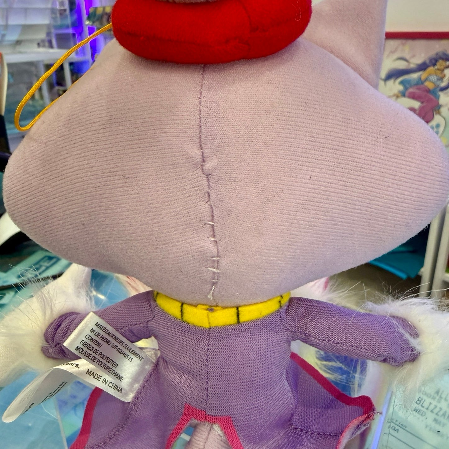 Close-up back view of a plush toy with a pale lavender body, red top, yellow collar, and white fluffy trim, photographed indoors with tags attached.