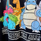Close-up of a black graphic T-shirt featuring colorful Pokémon characters including Venusaur, Charizard, and Blastoise, with white outlined text reading 'CATCH 'EM ALL!'.