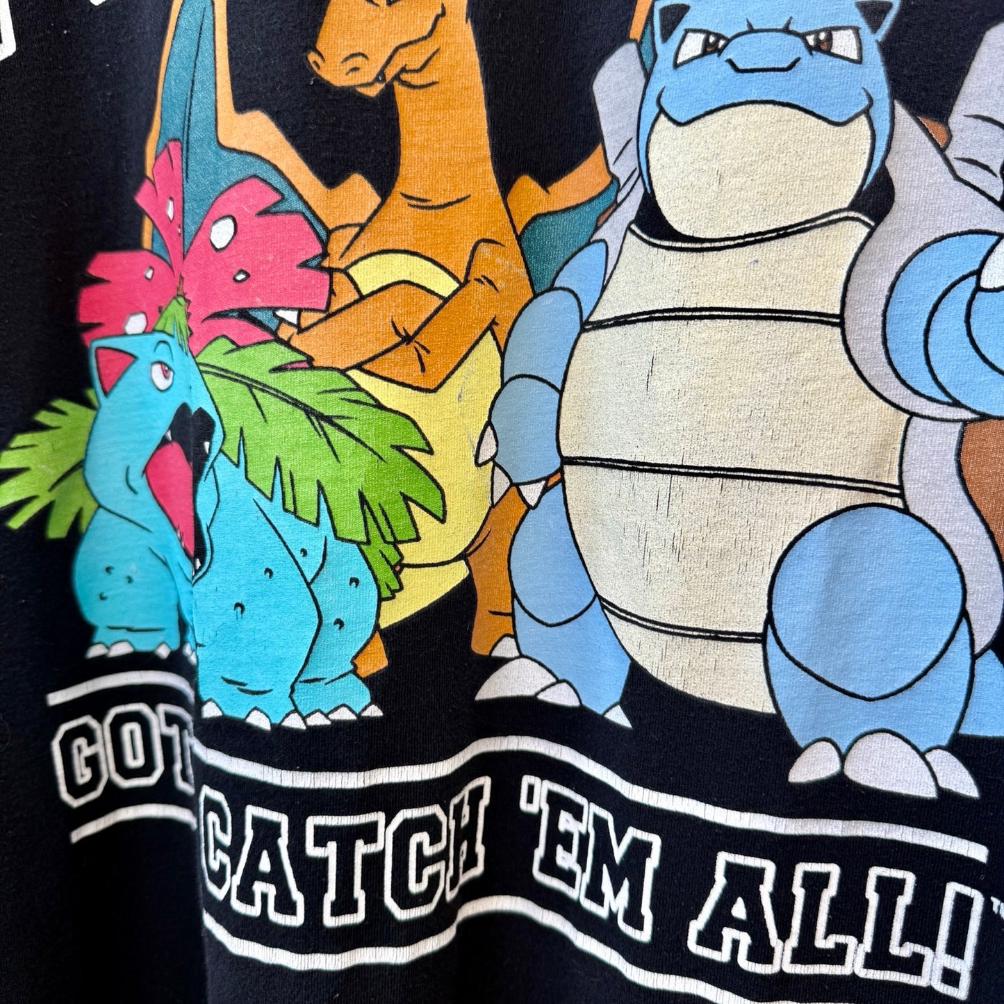 Close-up of a black graphic T-shirt featuring colorful Pokémon characters including Venusaur, Charizard, and Blastoise, with white outlined text reading 'CATCH 'EM ALL!'.