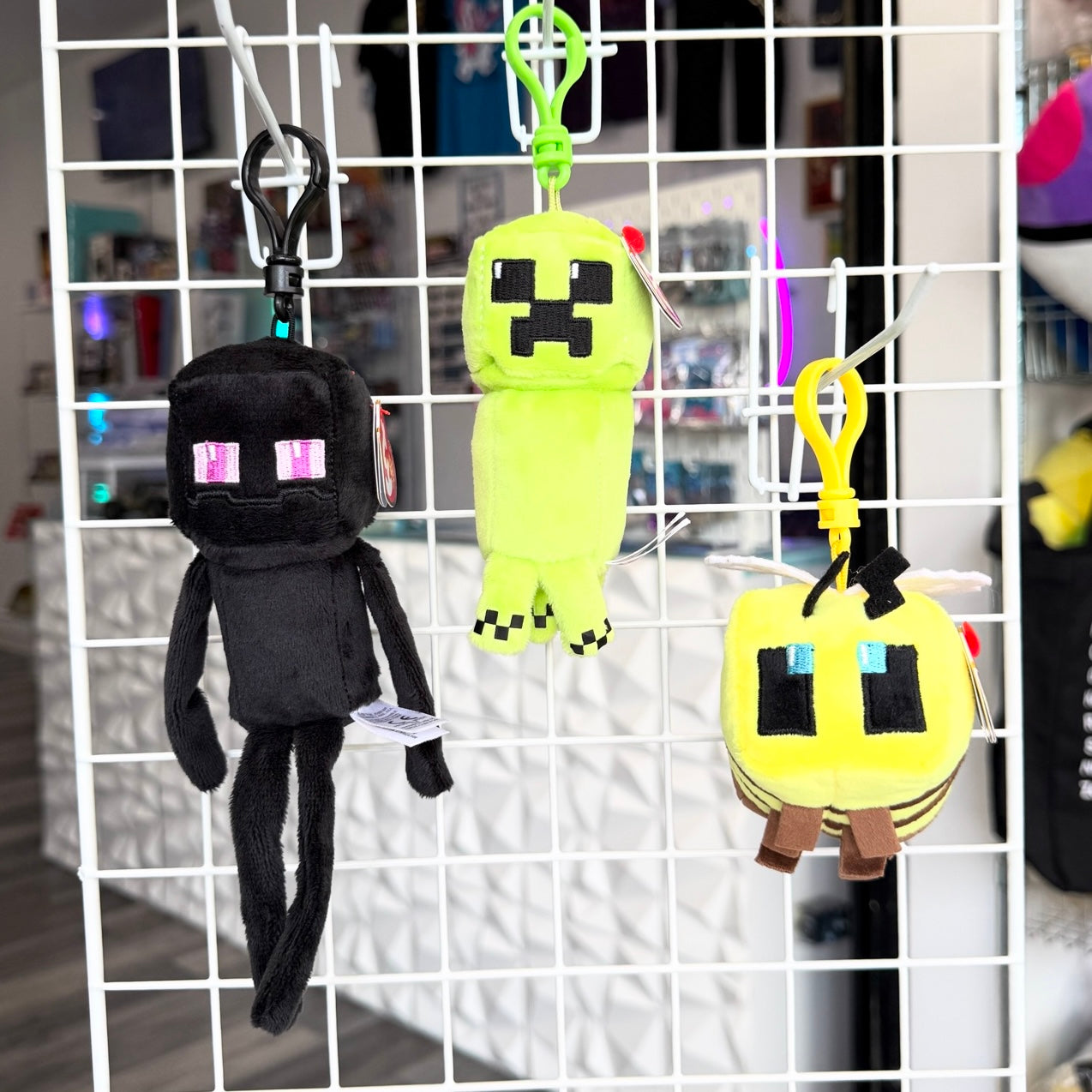 Three Minecraft-themed plush keychains hanging from a white wire display grid in a store. 