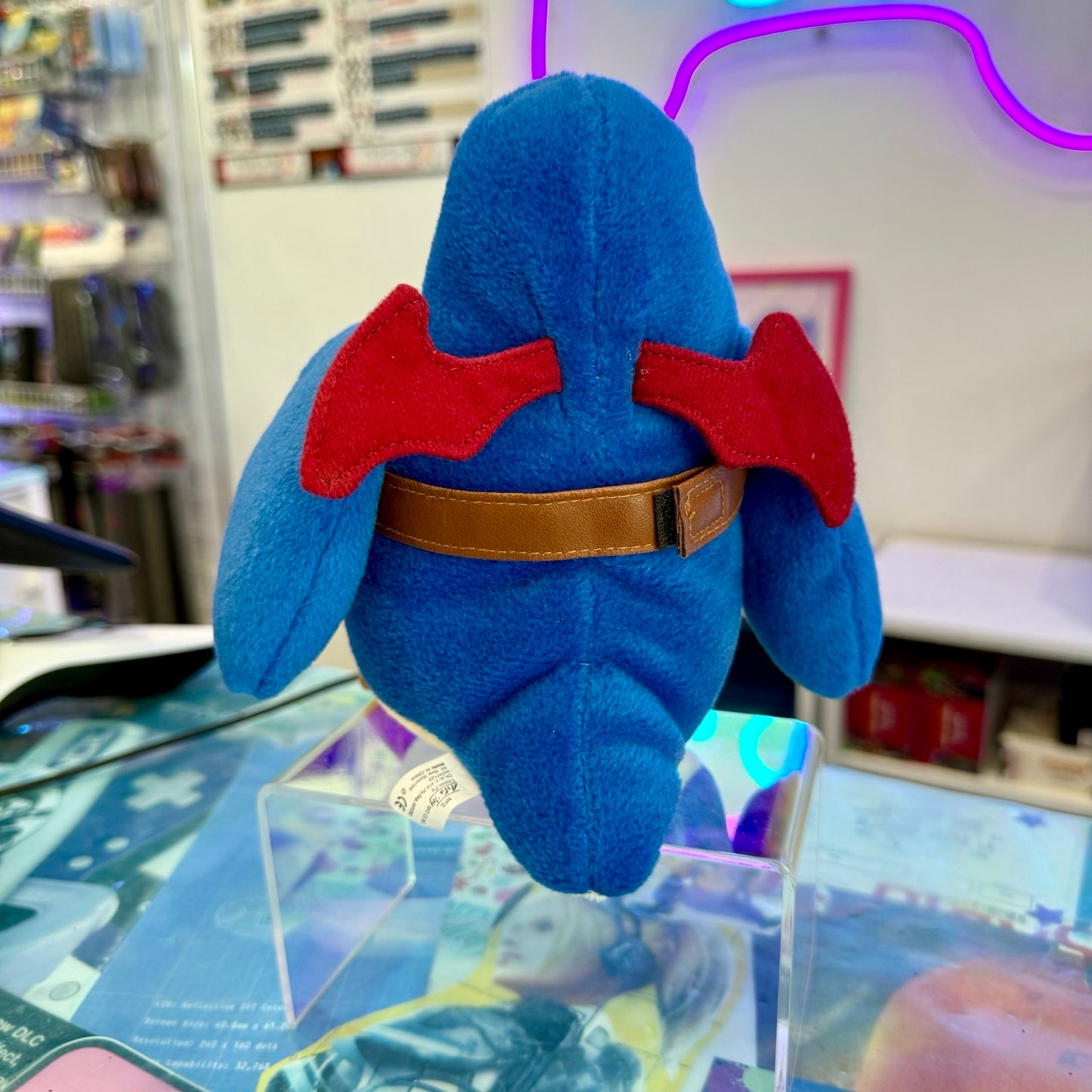 A blue plush toy with red wing-like accents and a brown belt, photographed from the back on a clear acrylic display stand in a retail store setting.