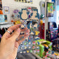 A hand holds a clear plastic sheet filled with multiple Pokémon Snorlax stickers photographed in a retail store with shelves and products blurred in the background.