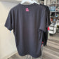 A dark charcoal gray t-shirt hanging on a white hanger, showing the back with a small pink graphic logo near the collar. The shirt is displayed in a retail store setting with other garments and shelves visible in the background.