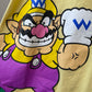 Close-up of a yellow T-shirt featuring a large printed graphic of Wario from Super Mario Bros., including his yellow hat, purple overalls, pink nose, and white chef hat with a blue W.