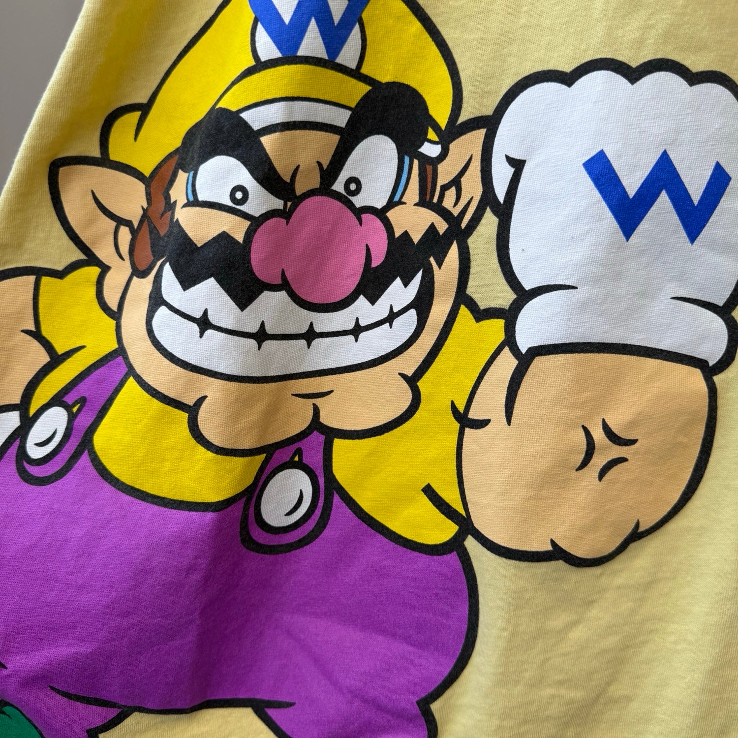 Close-up of a yellow T-shirt featuring a large printed graphic of Wario from Super Mario Bros., including his yellow hat, purple overalls, pink nose, and white chef hat with a blue W.