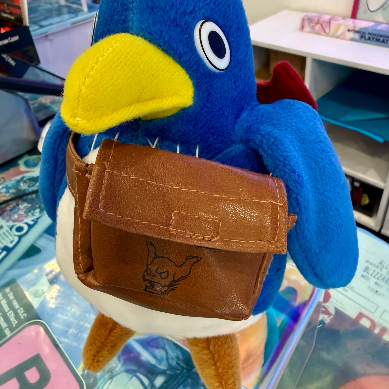 A blue plush bird character wearing a brown faux-leather pouch with a stitched flap and an embossed cartoon face, standing on a glass display counter in a retail setting.