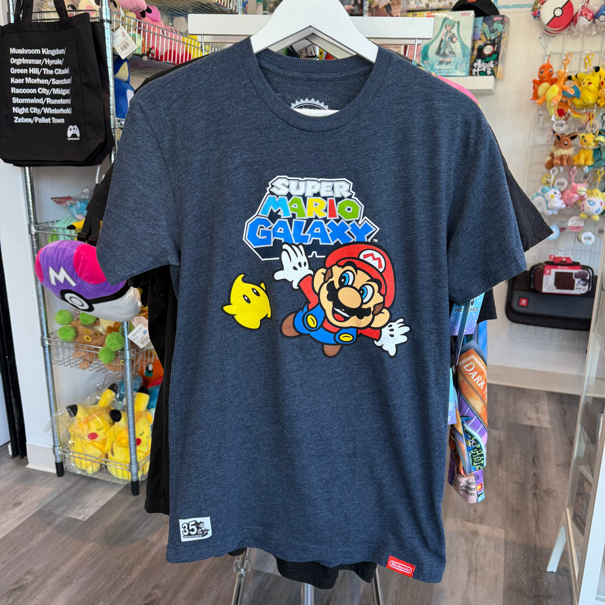 A dark heather blue t-shirt featuring a colorful 'Super Mario Galaxy' graphic on the front, hanging on a white hanger in a retail store display.