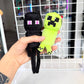 A hand holds two Minecraft plush toys, including a black Enderman and a green Creeper, in front of a white wire grid display.