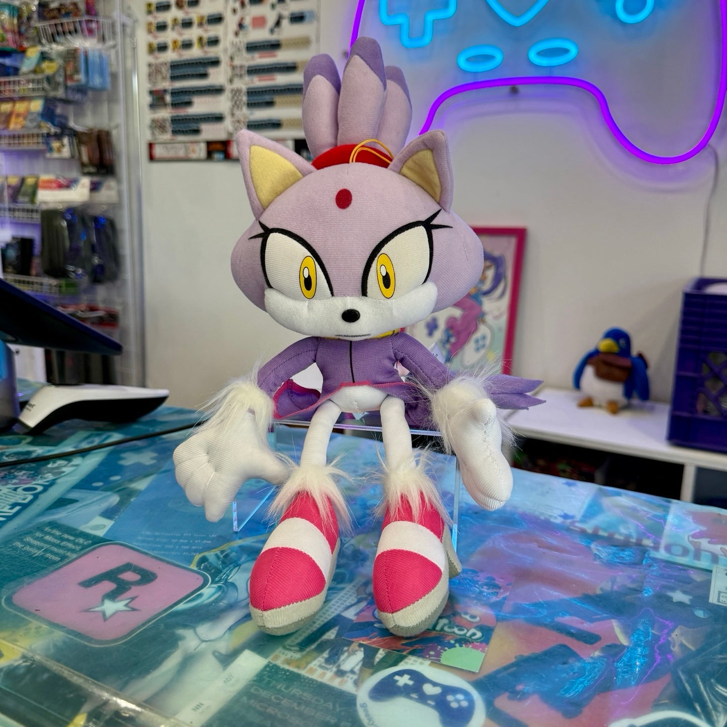 A plush toy of a purple cat character with yellow eyes, white gloves, and pink-and-white sneakers, displayed on a colorful gaming-themed surface in a retail setting.