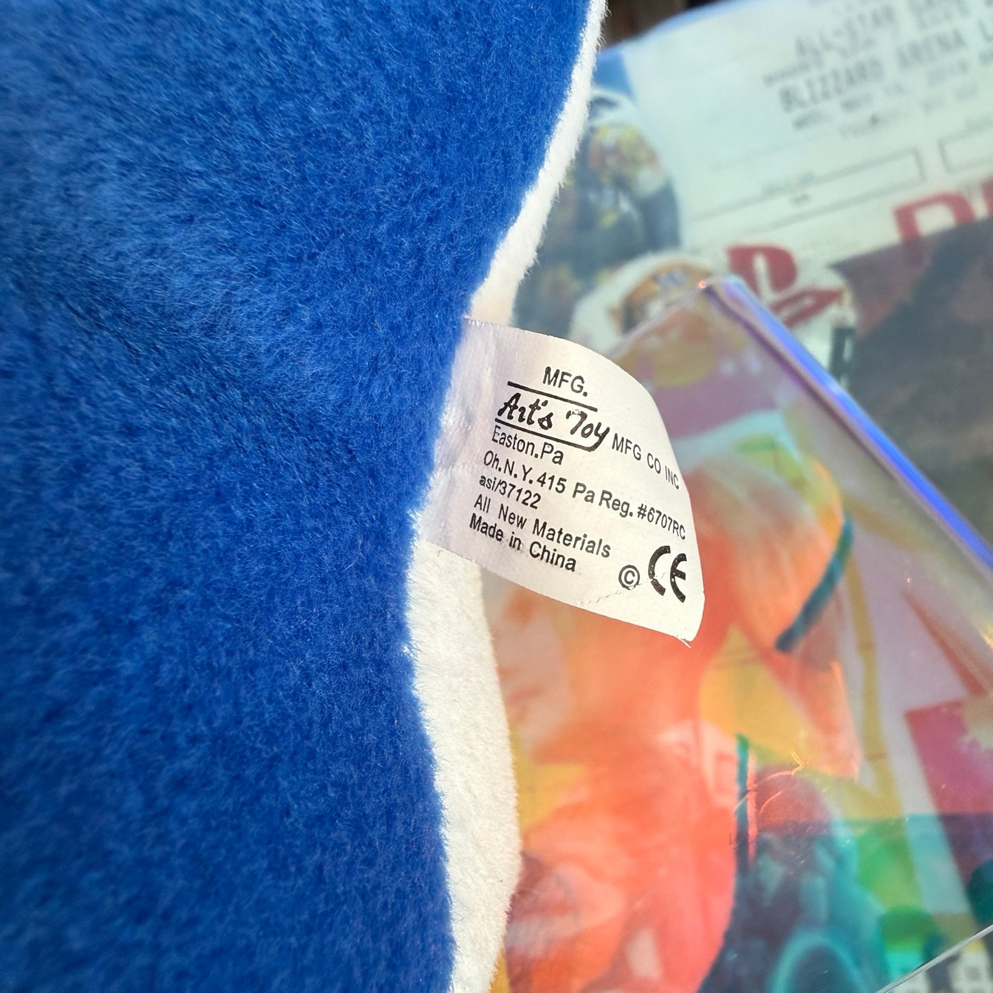 Close-up of a plush toy's white sewn-in tag reading 'MFG. Art's Toy MFG Co, PA' attached to a bright blue fuzzy fabric with a white border, with a blurred colorful background.