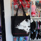 Black canvas tote bag with graphic from the video game, The World Ends With you on the front. Hanging on a metal shelf in a store setting.