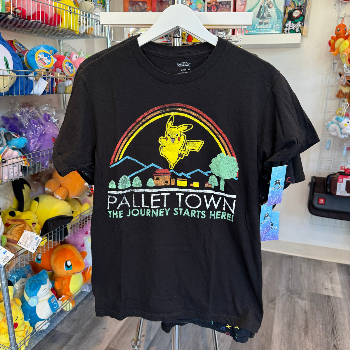 A black short-sleeve graphic t-shirt hanging on a white hanger in a retail store. The shirt features a colorful retro-style graphic of Pikachu under a rainbow with the text 'PALLET TOWN THE JOURNEY STARTS HERE!'.
