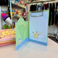 A green and blue Pokémon themed memo pad clipboard with a store in the background. 