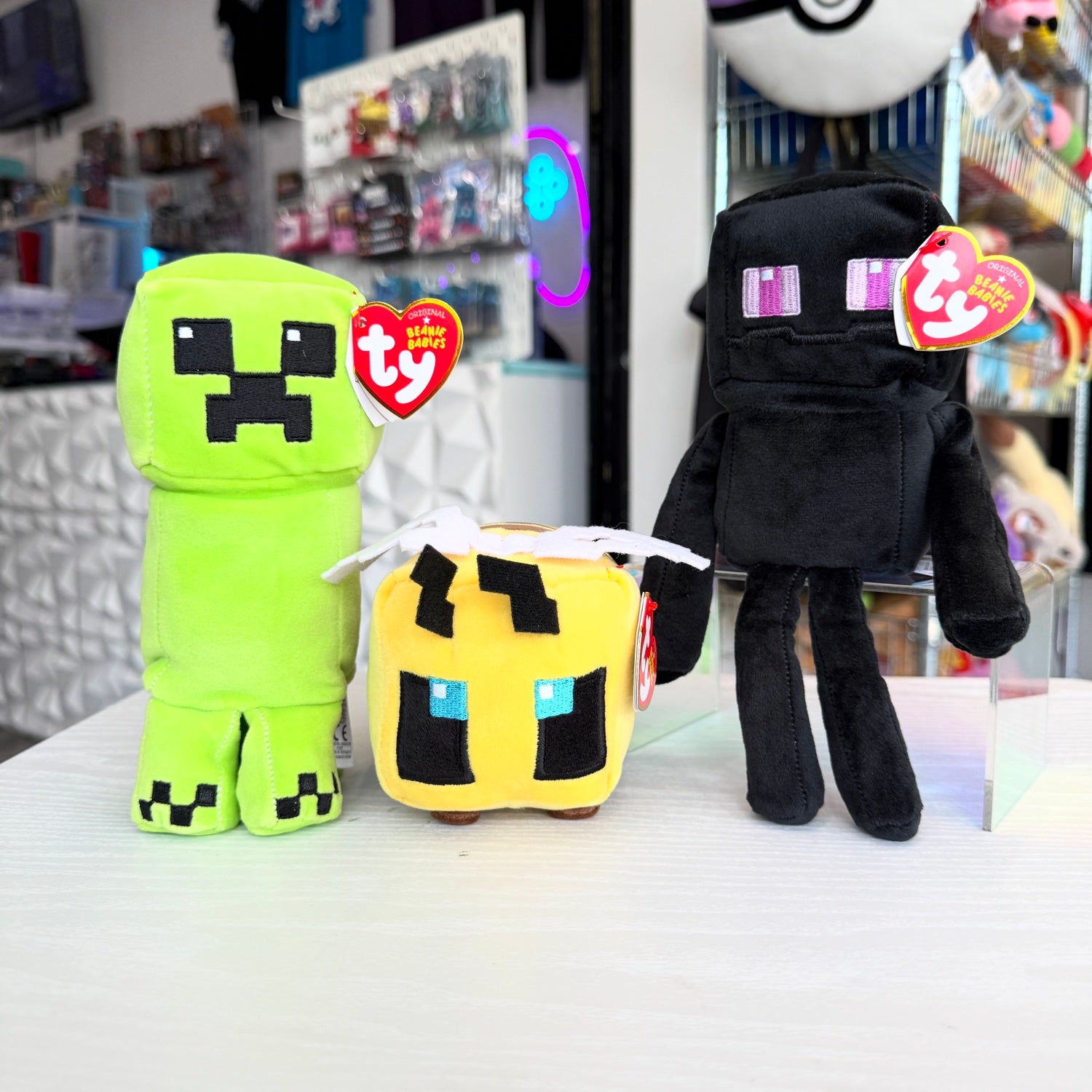 Three Minecraft plush toys from Ty brand, including a green Creeper, a yellow bee character, and a black Enderman, displayed on a white table with a blurred store background.