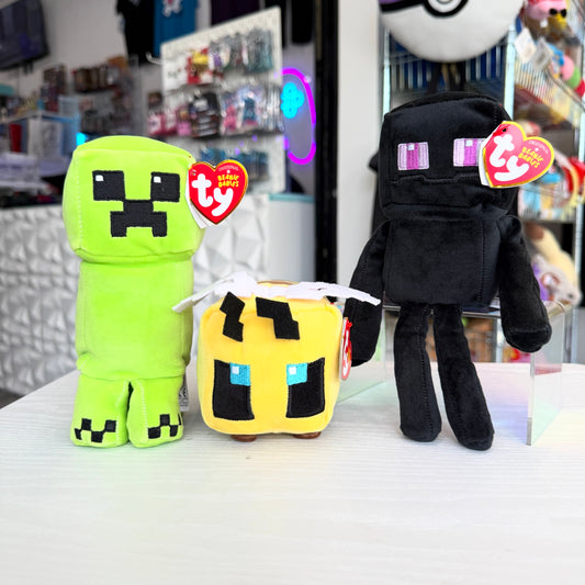 Three Minecraft plush toys from Ty brand, including a green Creeper, a yellow bee character, and a black Enderman, displayed on a white table with a blurred store background.